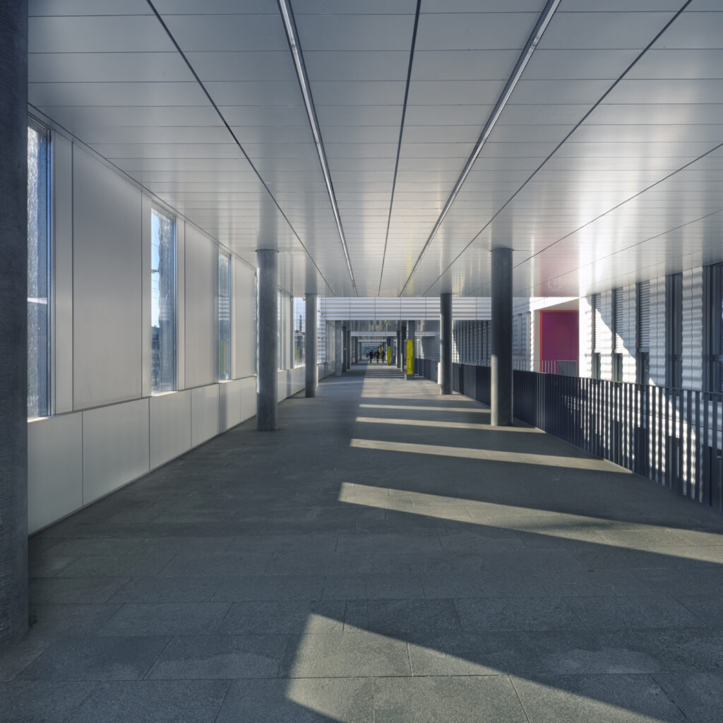 Architecture photography of the Jacob Burckhardt Haus in Basel with a covered walkway, repeating columns and geometric shadows on the floor. Architekturfotografie des Jacob Burckhardt Haus in Basel mit einem überdachten Steg, rhythmischen Stützen und geometrischen Schatten auf dem Boden.